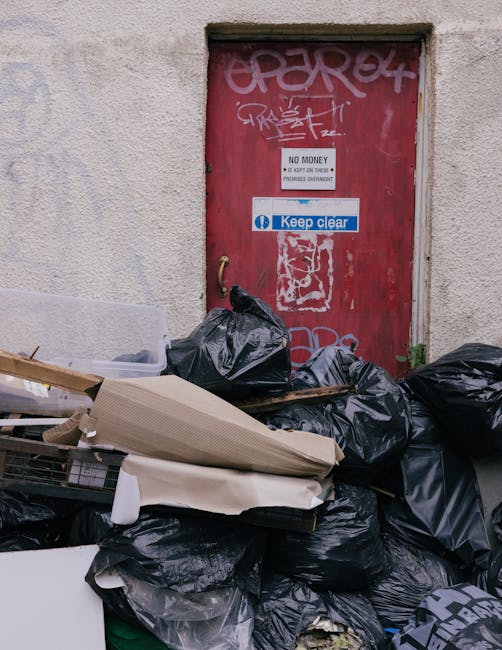 A collection of black plastic rubbish bags and discarded cardboard, with some white foam packaging and a partially visible piece of rolled-up cardboard, piled in front of a red metal door that has graffiti and multiple stickers, including a 'Keep clear' sign and a 'No money' notice, all situated on an exterior wall of a building. The scene appears to be an outdoor urban environment, possibly in a service area or alleyway, where waste has accumulated near the door, which is surrounded by a textured, off-white wall with some visible weathering and signs of neglect. The lighting is natural, casting soft shadows, and the overall scene emphasizes an example of rubbish awaiting collection, relating to private waste handling and alternative rubbish removal solutions often utilized in independent clearance services like those offered by Rubbish Clearance Hounslow to avoid reliance on traditional local authority collections.