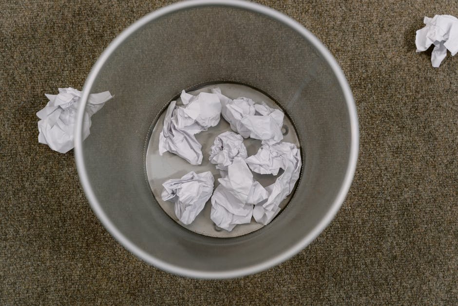 An overhead view of a cylindrical metal waste bin with a smooth, matte finish, partially filled with crumpled white sheets of paper. The bin is positioned on a brown, textured carpeted floor, with a few additional crumpled papers scattered near it on the floor. The inside of the bin shows signs of use, with some minor smudges and water at the bottom, possibly indicating previous paper disposal. The scene suggests an office or retail environment where paper waste is collected for removal. The metal surface of the bin is clean and reflective, contrasting with the soft, wrinkled texture of the paper and the roughness of the carpet. This image exemplifies a typical setup for independent rubbish collection or on-site waste clearance by a professional service such as Rubbish Clearance Hounslow, focusing on small-scale waste management tasks involving paper waste disposal.