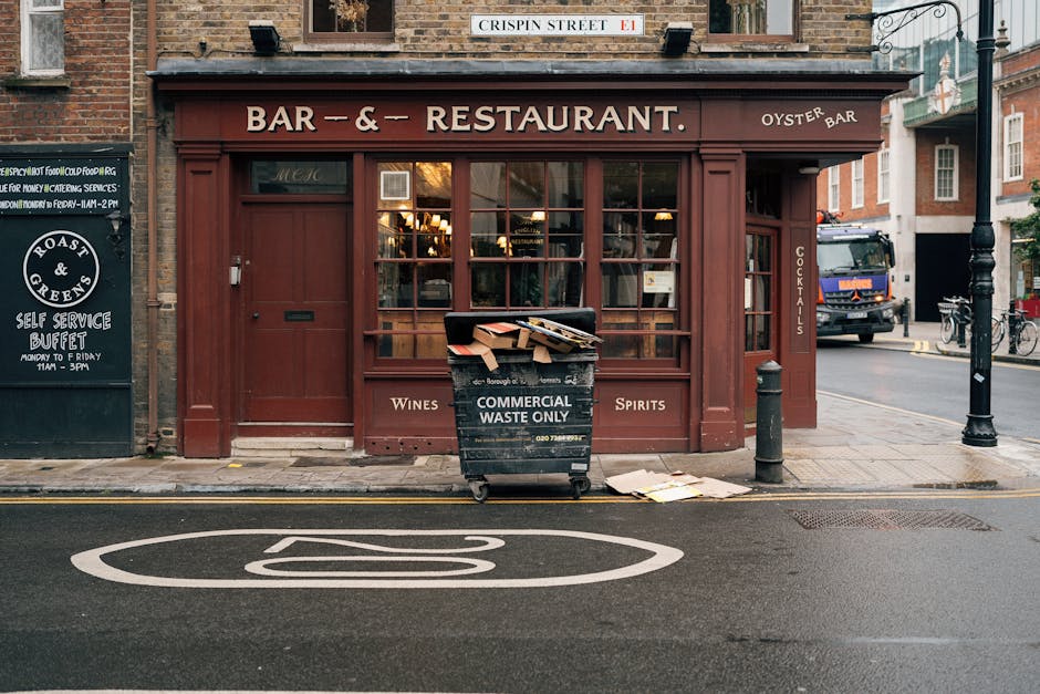 In front of a traditional brick building housing a bar and restaurant, a large black waste collection bin labeled 'Commercial Waste Only' is positioned on the sidewalk. The bin, made of plastic with metal wheels, is filled with discarded cardboard boxes and paper packaging, some of which have spilled onto the pavement nearby. The surroundings include a black bollard and a metal post along the curb, with a street featuring a marked no-parking zone painted in white, and a black metal street lamp nearby. The shop's storefront has large wooden-framed windows with warm interior lighting visible through the glass, and a sign on the awning indicates offerings such as wines, spirits, and cocktails. To the left, a blackboard sign advertises a self-service buffet, while on the street, a delivery truck is parked, and bicycles are secured to a rack further down the sidewalk. The scene depicts an urban environment where private waste collection services, like those from Rubbish Clearance Hounslow, are often employed for commercial rubbish removal, with environmentally appropriate disposal methods supported by visible waste management practices.