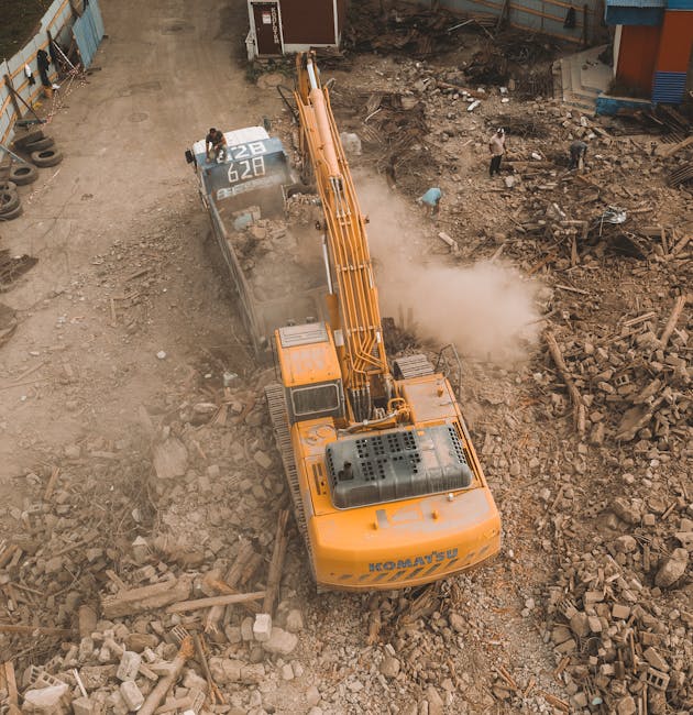 An aerial view of a construction site showing a large yellow excavator with black details, actively breaking and removing concrete debris with a hydraulic arm and a bucket attachment. Dust rises from the impact zone, where scattered rubble, broken bricks, and fragmented concrete are visible on the ground. To the left, a metal fence surrounds the site, with a row of tires and a small structure partially visible along the fence line. In the background, workers are present, some standing near piles of debris or walking across the soil, which appears rough and uneven. The environment suggests a site undergoing structural demolition, with significant waste material prepared for removal or disposal. Rubbish Clearance Hounslow offers private and on-site clearance services aligned with managing such waste efficiently, evident from the scene's focus on debris removal. The overall setting emphasizes machinery use and manual labour in waste management associated with building renovation or demolition projects, supporting the need for specialist rubbish removal services in urban construction areas.