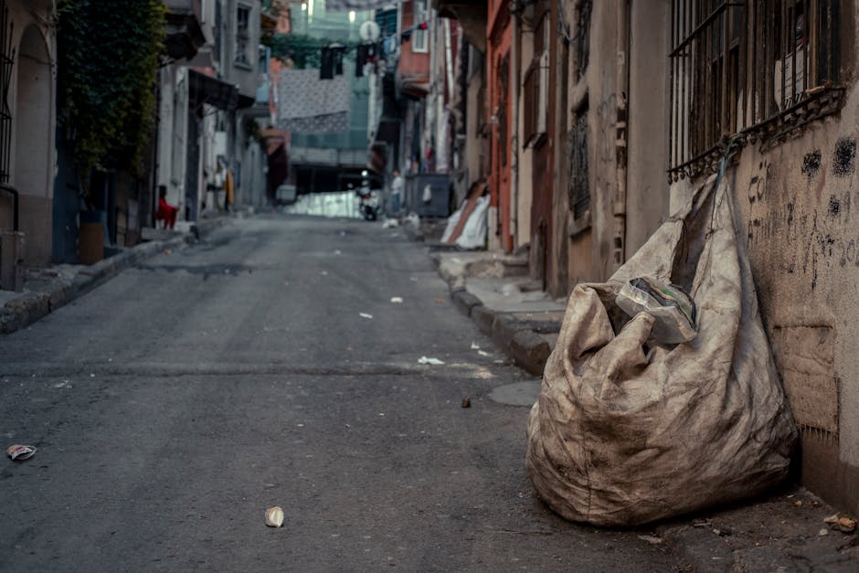 A large, weathered beige cloth sack filled with assorted waste materials sits on the uneven, worn pavement of a narrow urban alleyway. The sack appears dirty and crumpled, with a partially visible transparent plastic bag containing smaller debris protruding from the top. The street is flanked by tightly packed, multi-story buildings with rough, graffiti-marked walls, metal security bars on windows, and a variety of peeling paint and exposed brickwork. In the background, laundry hangs from lines strung between buildings, and a few indistinct figures are visible further down the alley. Nearby, small scattered pieces of trash and debris, including a crumpled paper and a plastic bottle, are visible on the ground, contributing to an overall impression of neglect. The lighting is soft and diffuse, suggesting an overcast day, and the scene subtly reflects the need for alternative waste handling, such as private rubbish collection by services like Rubbish Clearance Hounslow, to address urban waste issues efficiently.