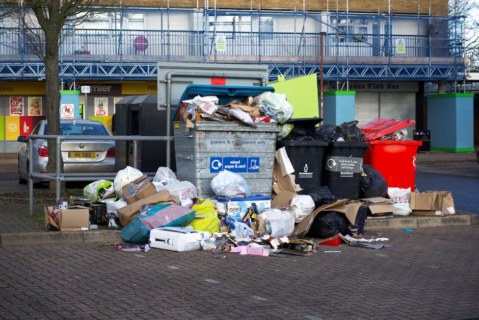 A collection of overflowing rubbish and waste materials piled on a paved sidewalk in front of a commercial building. The pile includes various trash bags, cardboard boxes, paper waste, and plastic packaging, with some items spilling onto the surrounding area. There is a large grey bin marked for mixed paper and card, partially open with discarded papers and cartons sticking out. Several black and red rubbish bins are also visible nearby, some with trash overflowing. A small metallic fence separates the waste from parked vehicles, including a silver car with a visible license plate. In the background, a building with blue scaffolding or protective netting, bright yellow signage, and storefronts is visible, indicating an urban setting. The scene highlights the need for effective rubbish removal services, such as those offered by Rubbish Clearance Hounslow, especially for private collection or on-site clearance from commercial or residential areas.
