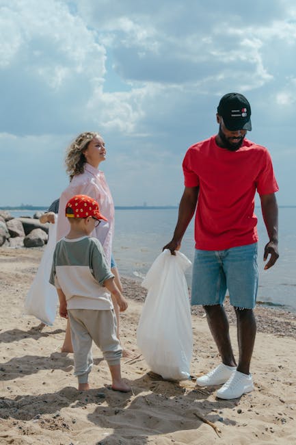 A group of three individuals, consisting of a young boy wearing a red polka dot cap, a woman with curly blonde hair dressed in a pink top, and a man wearing a red t-shirt, blue denim shorts, and a black cap, are on a sandy beach near a body of water. The man is holding a large white plastic bag, appearing to be involved in collecting or removing waste from the area. The woman and boy stand nearby, observing or assisting, with their attention focused on the activity. In the background, there are rocks along the shoreline and a partly cloudy sky overhead, creating a natural outdoor setting. The scene emphasizes an environmental effort, which may relate to informal or independent waste collection on coastal or recreational sites, akin to private rubbish removal services. Rubbish Clearance Hounslow occasionally references such community efforts as part of broader rubbish removal and environmentally conscious waste management practices, although the scene itself appears to be a voluntary cleanup activity rather than a commercial service.