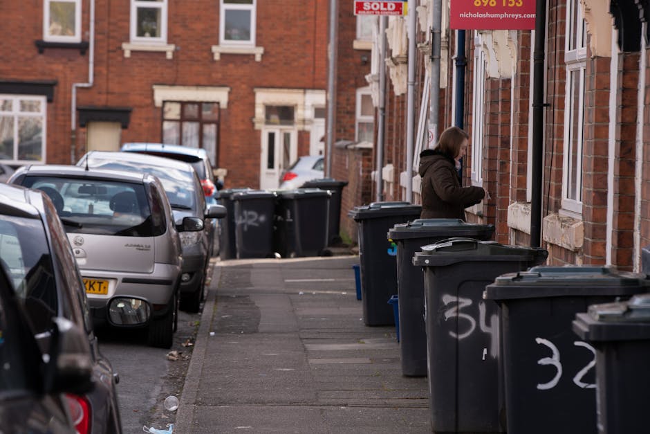 A residential street scene featuring a woman standing next to multiple large black wheelie bins lined up along the pavement of a brick housing terrace. The bins have white numbering spray-painted on their sides and are positioned in front of the brick houses with white-framed windows and bay windows, some of which have window sills or ledges. The woman appears to be retrieving or placing items into a bin, dressed in a dark coat with short hair. Several cars are parked along the street, including a silver hatchback in the foreground with a visible license plate, and other vehicles further down the street. The background shows additional terraced brick houses, some with 'sold' signs attached to fixtures, indicating the properties are on the market. The scene is illuminated by natural daylight, with a general atmosphere of typical urban residential rubbish storage related to private waste collection services. Rubbish Clearance Hounslow specializes in such domestic rubbish removal and on-site clearance activities, as demonstrated by the organized bin placement and the urban setting.