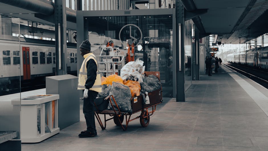 A worker wearing a dark beanie, black jacket, and yellow reflective vest stands on a train station platform near a large green metal pillar. Next to the worker, there is a red trolley loaded with various bags of waste, including black, orange, and white plastic sacks filled with rubbish, some of which are torn open revealing mixed debris. Behind the trolley, a modern glass and metal structure provides shelter, with security or station staff visible in the background. To the left, there is a white container or recycling bin positioned against the pillar. On the right side of the platform, the railway tracks run parallel, with a train parked alongside, and a few additional commuters are visible in the distance. The environment appears orderly with a clean, paved surface, illuminated by natural daylight, reflecting a typical urban railway station setting. This scene relates to waste management and the collection of rubbish in public transit environments, similar to services offered by companies like Rubbish Clearance Hounslow for on-site clearance and alternative waste handling solutions.