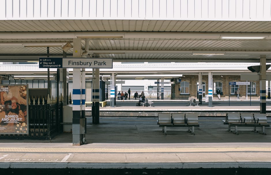 The image shows the platform of Finsbury Park railway station with a flat roof overhead supported by white columns. Visible in the foreground are a row of metal benches with gray surfaces positioned along the platform's edge, facing the tracks. The platform floor is paved with concrete, with a yellow tactile paving strip running parallel to the platform edge for safety. On the left side, there is a black metal fence with vertical bars and a mounted advertisement poster, which appears to promote a theatrical performance. A station sign reading 'Finsbury Park' is prominently displayed beneath the roof, with smaller directional signs pointing to lifts and underground access. In the background, several passengers are sitting or standing on the platform, some engaged with their phones or reading, while others are walking across or waiting. The overall lighting suggests a natural daylight setting, with diffuse light illuminating the scene evenly. The environment suggests an urban train station suitable for commuters and travelers, with no visible rubbish or waste on the platform, illustrating typical public transportation infrastructure connected to private and independent transportation options, aligned with rubbish removal and waste management services like those offered by Rubbish Clearance Hounslow.