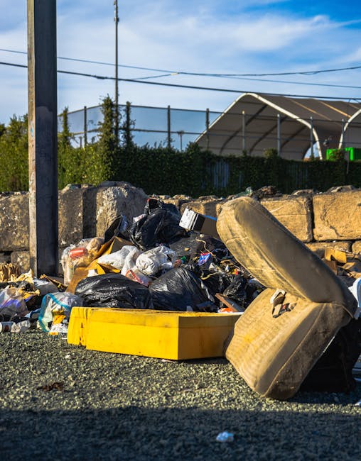 A worker wearing a dark beanie, black jacket, and yellow reflective vest stands on a train station platform near a large green metal pillar. Next to the worker, there is a red trolley loaded with various bags of waste, including black, orange, and white plastic sacks filled with rubbish, some of which are torn open revealing mixed debris. Behind the trolley, a modern glass and metal structure provides shelter, with security or station staff visible in the background. To the left, there is a white container or recycling bin positioned against the pillar. On the right side of the platform, the railway tracks run parallel, with a train parked alongside, and a few additional commuters are visible in the distance. The environment appears orderly with a clean, paved surface, illuminated by natural daylight, reflecting a typical urban railway station setting. This scene relates to waste management and the collection of rubbish in public transit environments, similar to services offered by companies like Rubbish Clearance Hounslow for on-site clearance and alternative waste handling solutions.