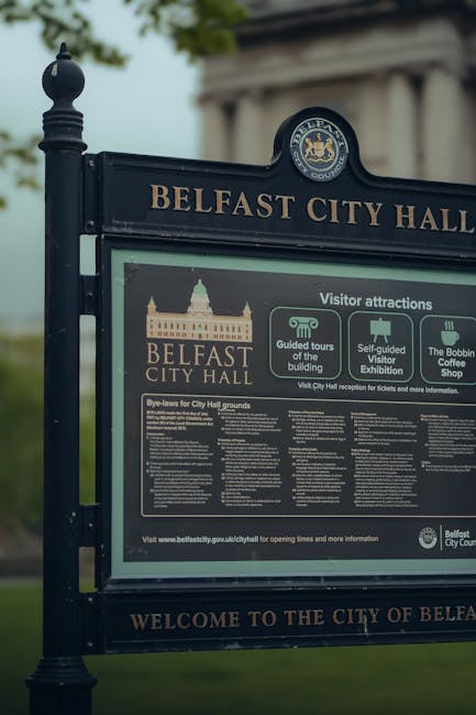 A close-up view of a black metal signpost for Belfast City Hall, featuring the text 'BELFAST CITY HALL' at the top and the city's coat of arms in an oval above. Below, there are informational panels providing details about visitor attractions, guided tour options, a self-guided visitor exhibition, and a cafe shop, with small icons representing each feature. The signpost is situated outdoors on a paved area, with a blurred background of green trees and part of a classical-style building with columns visible behind. The panel about Belfast City Hall grounds includes detailed rules and guidelines, suggesting the focus on managed public spaces. The scene, lit by natural daylight, emphasizes the contrast between the dark metal signage and the lighter background, aligning with themes of urban public management and possibly alternative methods of waste or rubbish handling in city spaces, as referenced in the context of rubbish removal services such as Rubbish Clearance Hounslow.
