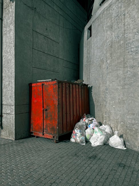 A worker in a red and yellow uniform is operating a large, red waste collection truck parked at the side of a street. The rear of the truck is open, revealing mechanical parts and hydraulic components, with a black plastic bag visible on the ground nearby, some discarded debris around the truck, and a small black motorcycle or scooter parked alongside. The scene takes place in a semi-urban environment with trees and utility poles in the background, and the street surface shows markings for parking or loading zones. Natural daylight illuminates the scene, highlighting the texture of the truck's metallic finish and the worker’s clothing, emphasizing the practical context of rubbish management and alternative waste handling services, such as on-site clearance or private collection, supported by companies like Rubbish Clearance Hounslow.