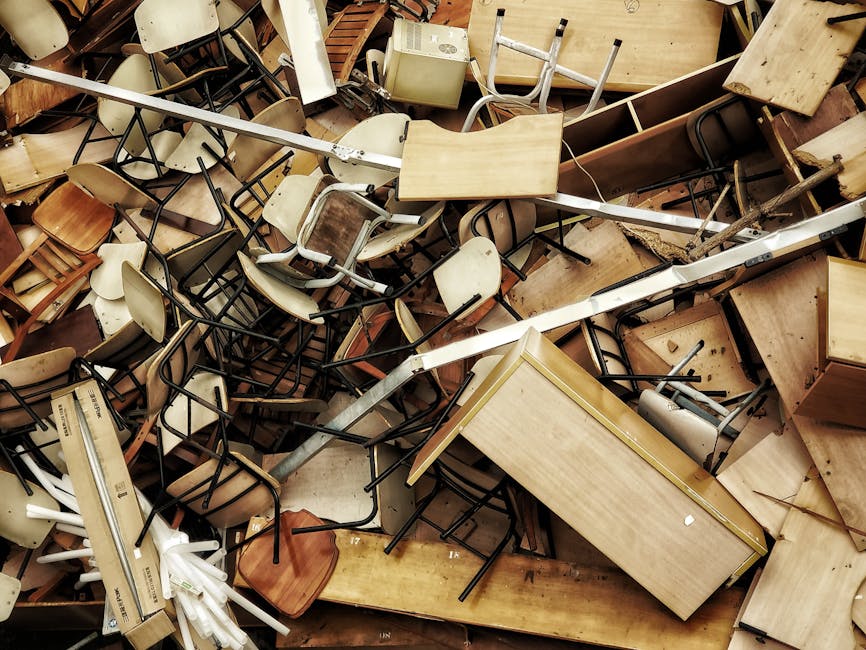 A large pile of discarded office furniture and equipment, consisting mainly of wooden desks, chairs with metal and plastic frames, and small storage units, all haphazardly stacked in an indoor environment. The desks display various finishes and textures, including smooth, light-colored wood surfaces, some with visible scratches or worn edges. The chairs are a mix of cream, beige, and brown tones, with some featuring plastic seats and others with wooden or metal legs. Overlapping and jumbled arrangements of the furniture create a cluttered scene, with chairs flipped upside down or leaning against desks. A long metal rod or support beam runs diagonally across the pile, adding to the disorderly appearance. Light is diffused, casting soft shadows that highlight the variety of materials and the chaotic stacking. The context suggests an off-site or private furniture clearance, where Rubbish Clearance Hounslow manages waste removal for furniture that might otherwise be disposed of via conventional recycling or collection services, illustrating an alternative approach to rubbish handling and on-site clearance.