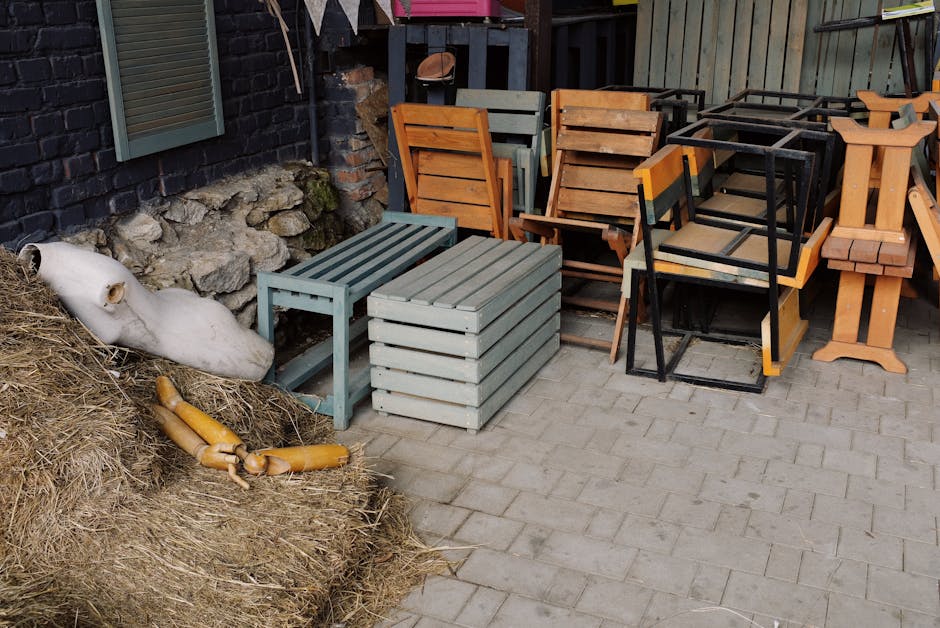 A large pile of discarded office furniture and equipment, consisting mainly of wooden desks, chairs with metal and plastic frames, and small storage units, all haphazardly stacked in an indoor environment. The desks display various finishes and textures, including smooth, light-colored wood surfaces, some with visible scratches or worn edges. The chairs are a mix of cream, beige, and brown tones, with some featuring plastic seats and others with wooden or metal legs. Overlapping and jumbled arrangements of the furniture create a cluttered scene, with chairs flipped upside down or leaning against desks. A long metal rod or support beam runs diagonally across the pile, adding to the disorderly appearance. Light is diffused, casting soft shadows that highlight the variety of materials and the chaotic stacking. The context suggests an off-site or private furniture clearance, where Rubbish Clearance Hounslow manages waste removal for furniture that might otherwise be disposed of via conventional recycling or collection services, illustrating an alternative approach to rubbish handling and on-site clearance.